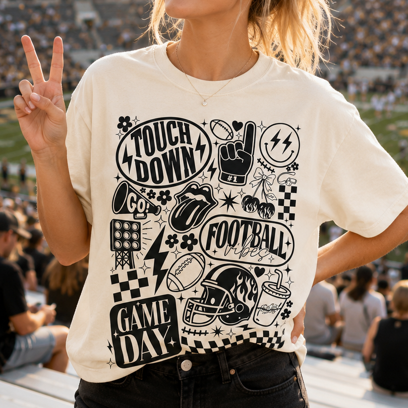A woman wearing a graphic t-shirt stands in a stadium, making a peace sign. The t-shirt is off-white with a black graphic print featuring football-themed illustrations and the words 'TOUCH DOWN', 'GAME DAY', and 'TOUCH IT UP'.